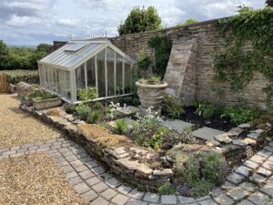 Classic garden design with a greenhouse, raised bed, and dry-stacked stone walls installed by landscapers in Derbyshire.