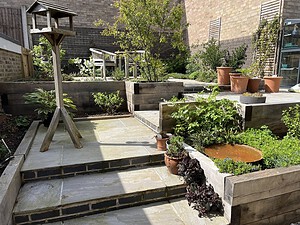 Multi-tiered garden design showcasing natural stone steps, terraced wooden sleeper planters filled with lush green plants, and a rustic bird table