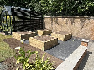 Four large timber raised vegetable beds on a dark grey block-paved path in a dedicated kitchen garden layout next to a greenhouse